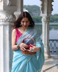 Woman in a light blue silk cotton saree with a pink blouse, holding a woven basket, standing against an architectural background.