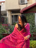 Woman in a pink saree standing outdoors with greenery and a building in the background