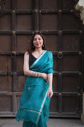 Woman in a teal saree standing against a textured brown wall.
