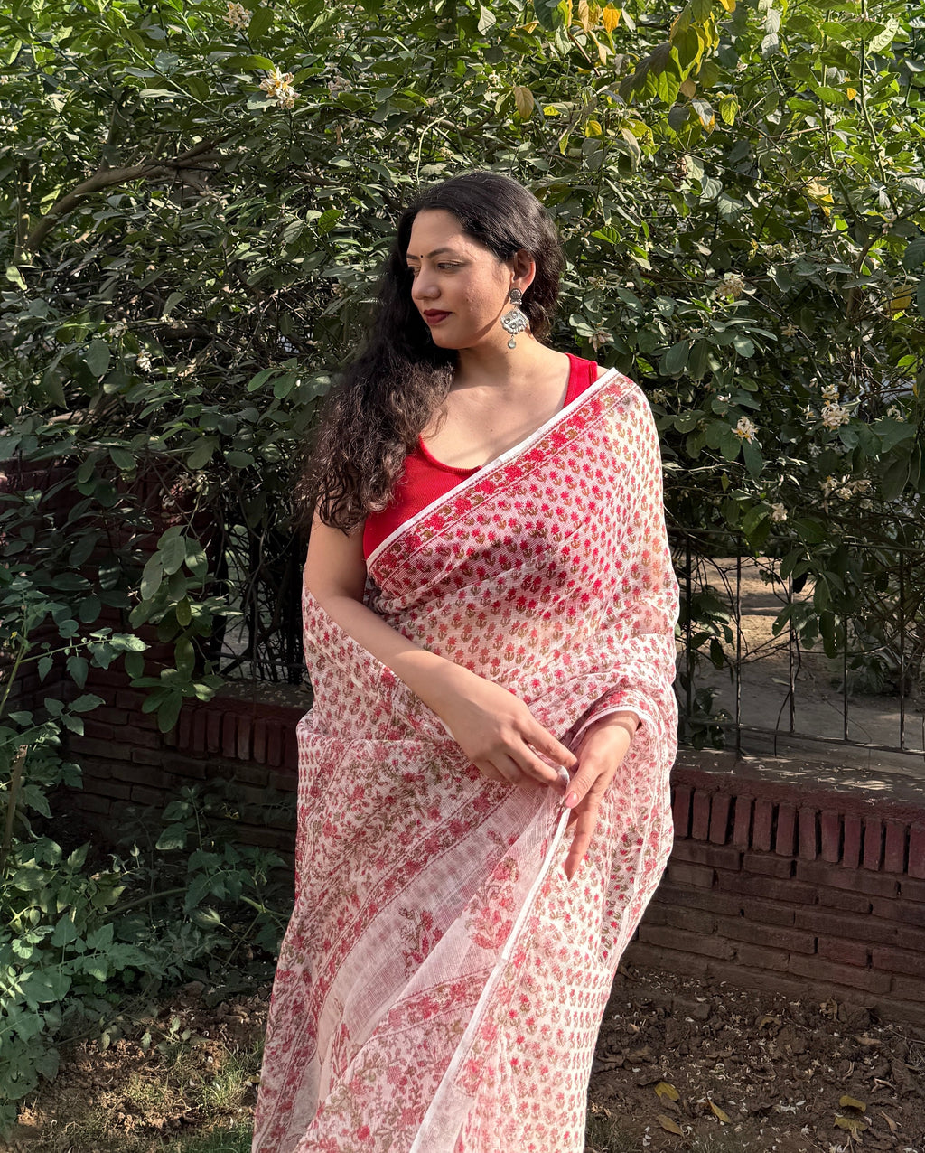 Woman wearing a red and white kota doria cotton patterned saree standing in front of green foliage.