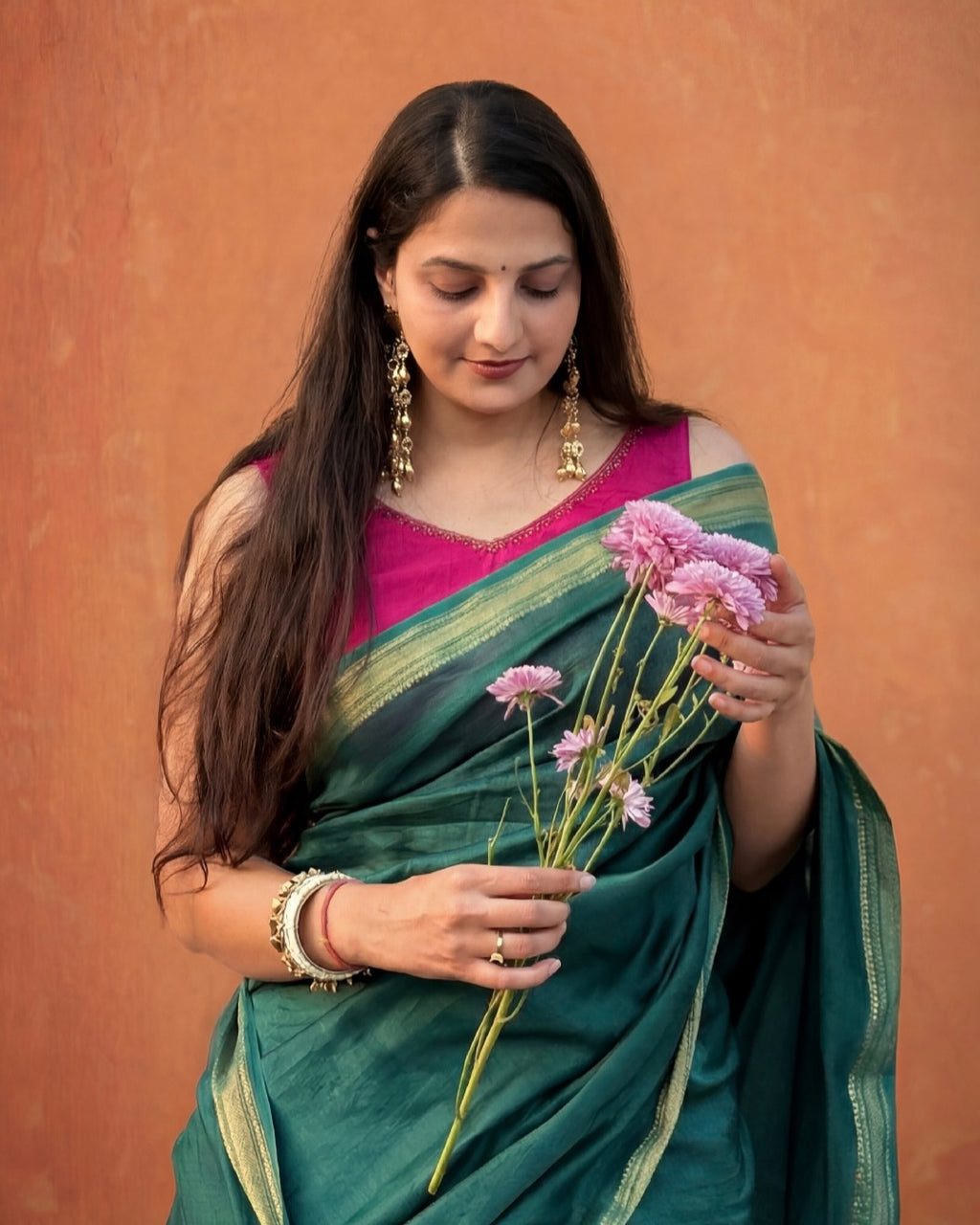 Woman in a green silk cotton saree with golden border holding pink flowers against a brown background